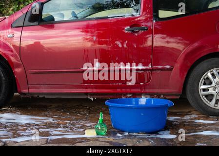 Durante un lavaggio manuale da un'auto rossa fluisce acqua, evidenziando un secchio blu e i prodotti per la pulizia su un vialetto di sapone Foto Stock