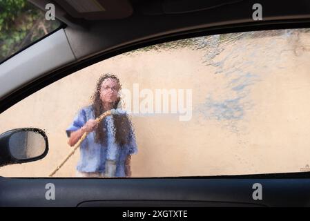 Vista dall'interno di un'auto che guarda fuori attraverso una finestra a vapore, mostrando una donna anonima che lava l'auto. Sembra concentrata sul suo compito, non guardando t Foto Stock