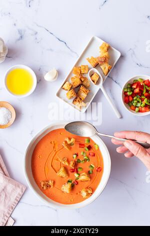 Vista dall'alto della tradizionale zuppa spagnola di gazpacho di pomodoro, adornata con crostini e verdure a dadini su una ciotola bianca, una persona irriconoscibile che tiene in mano Foto Stock
