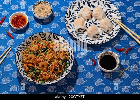 Dall'alto, una vivace esposizione di cibo cinese su un tavolo floreale blu con spaghetti di gamberi, gnocchi al vapore in una ciotola a motivi geometrici e condimenti lik Foto Stock