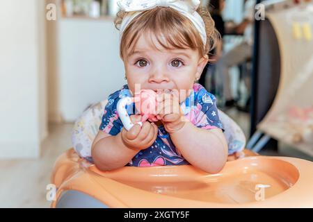 Una bambina simpatica si siede su un seggiolone a casa, masticando giocosamente un giocattolo colorato. I suoi occhi curiosi e il suo abbigliamento casual creano una scena affascinante Foto Stock