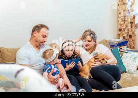 Un'accogliente scena familiare in cui due adulti e due bambini, insieme al loro cane, si incontrano su un divano, leggono e giocano insieme Foto Stock