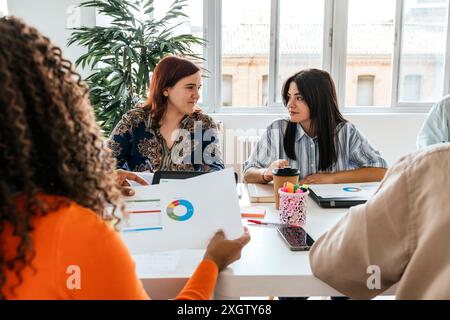 Un gruppo multietnico di professionisti collabora ad un tavolo disordinato in uno spazio di coworking ben illuminato. Due donne si impegnano attivamente con gli occhi l'una sull'altra, Foto Stock