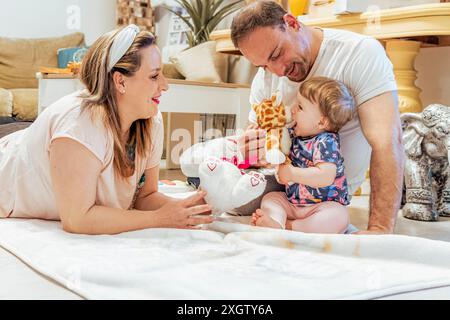 Un momento di gioia in famiglia catturato mentre i genitori giocano con il loro bambino su un accogliente pavimento del soggiorno. Il padre tiene in mano una giraffa giocattolo mentre il bambino si allontana Foto Stock