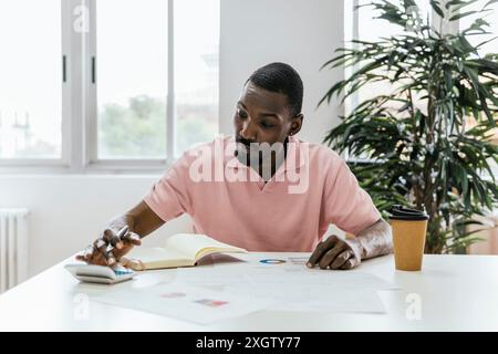 Uomo afroamericano che si occupa di carte aziendali e documenti in uno spazio di coworking, sembra concentrato e determinato. Guarda in basso con attenzione, mare Foto Stock