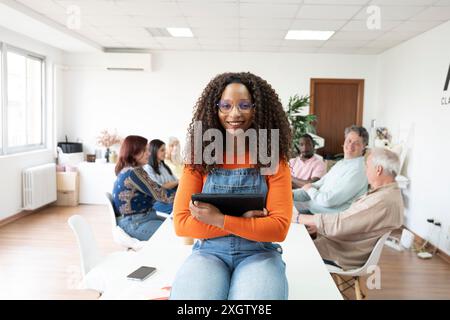 Donna afroamericana che guarda la macchina fotografica, tiene il tablet in uno spazio di coworking diversificato con persone di varie età ed etnie impegnate in di casuali Foto Stock