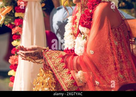 Vista corta e irriconoscibile delle mani di una sposa adornata da intricati disegni all'henné durante una cerimonia di nozze indiana, completata dal suo rosso vibrante Foto Stock