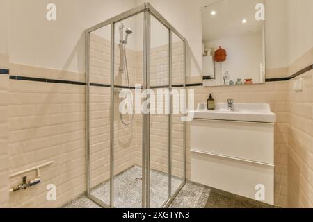 Un bagno contemporaneo con piastrelle in ceramica beige, box doccia ad angolo in vetro e lavabo bianco a parete che si riflette su un grande specchio. Foto Stock