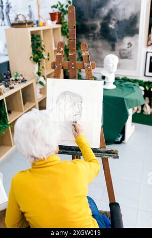 Anonima donna anziana con i capelli grigi, che indossa un maglione giallo, si concentra sulla pittura di un ritratto su un cavalletto in uno studio d'arte ben illuminato pieno di Foto Stock