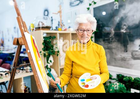 Una donna anziana allegra con i capelli bianchi dipinge su una tela in uno studio d'arte illuminato, tenendo in mano una tavolozza e una spazzola Foto Stock