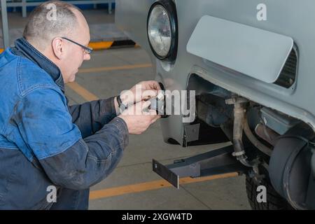 Un meccanico con abbigliamento da lavoro blu installa un gancio di traino su una vettura bianca in un'officina automobilistica Foto Stock