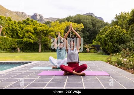 La coppia birazziale pratica yoga in un giardino sereno, con spazio per copiare. Si divertono insieme a una tranquilla sessione di allenamento all'aperto. Foto Stock