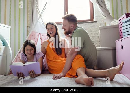 La famiglia di tre persone ama leggere un libro insieme sul pavimento di una sala giochi Foto Stock
