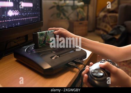 Primo piano di un bambino irriconoscibile che inserisce la cartuccia nella console d'epoca tenendo in mano il controller con un gioco vecchio stile visibile sullo schermo della TV Foto Stock