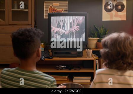 Vista posteriore di due bambini seduti sul pavimento a guardare la TV che proietta film horror sugli zombie in un accogliente soggiorno rétro con piante e poster a parete e una console di gioco in vecchio stile sotto lo schermo TV Foto Stock