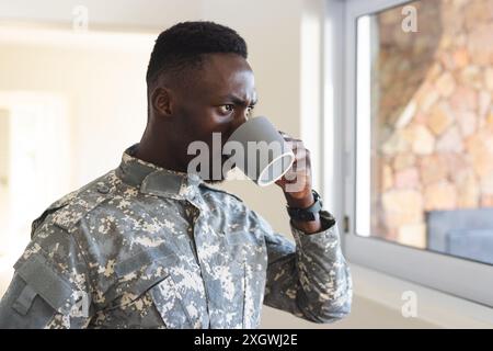 Premuroso soldato afroamericano a casa che tiene il caffè e guarda fuori dalla finestra. Depressione, ptsd, servizio militare, esercito, ritorno, a casa, uomini Foto Stock