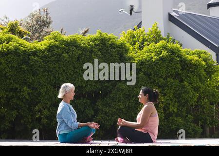 Due amiche anziane felici e diverse che praticano la meditazione yoga sedute in giardino, spazio fotocopie. Posizione Lotus, yoga, benessere, meditazione, amici Foto Stock