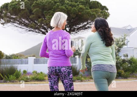 Vista posteriore di due amiche anziane felici e diverse che camminano per fare esercizio fisico sulla strada di campagna, parlando. Amicizia, fitness, esercizio fisico, attività e salute Foto Stock