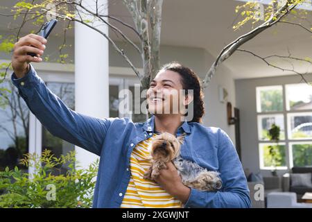 Il giovane uomo birazziale si fa un selfie all'aperto con il suo cane. Sta catturando un momento di gioia in un giardino di casa. Foto Stock