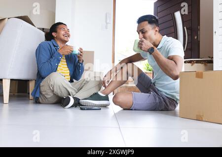 Giovane uomo birazziale e uomo mediorientale, una coppia gay, si godono una pausa caffè a casa. Condividono una risata mentre sono seduti sul pavimento in mezzo a una scatola in movimento Foto Stock