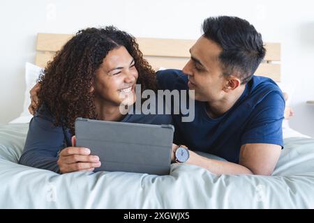 La coppia birazziale gay ama trascorrere il tempo libero a casa. Condividono un momento di gioia guardando un tablet nella loro camera da letto. Foto Stock