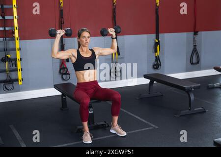 Vesti la giovane donna caucasica che solleva manubri in palestra. È concentrata sull'allenamento di forza, seduta con attrezzature per esercizi in background. Foto Stock