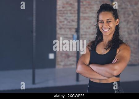 Una giovane donna birazziale forte e in forma sorride con fiducia in palestra, con spazio copia inalterato. Il suo abbigliamento atletico suggerisce un impegno per il fitness a. Foto Stock
