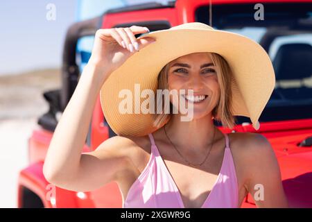 Una giovane donna caucasica indossa un costume da bagno rosa all'aperto durante un viaggio in auto. Il suo cappello a tesa larga aggiunge un tocco di eleganza al suo look da spiaggia. Foto Stock