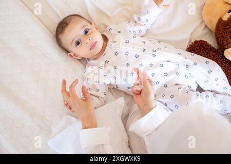 Bambino in pigiama a forma di stella disteso su un letto bianco con un orsacchiotto, assistito da un genitore in hijab. Vista dall'alto, primo piano, perfetta per la famiglia e i genitori Foto Stock