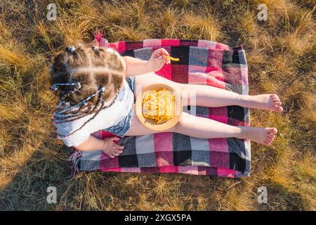 Una ragazza in bicchieri blu siede su una coperta in un prato con erba secca e mangia patatine fritte (vista dall'alto). Soleggiata giornata estiva in campagna. Picnic all'aperto Foto Stock