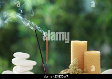 Bastoncini di incenso che si muovono vicino a pietre, candele e fiori secchi su sfondo verde sfocato Foto Stock