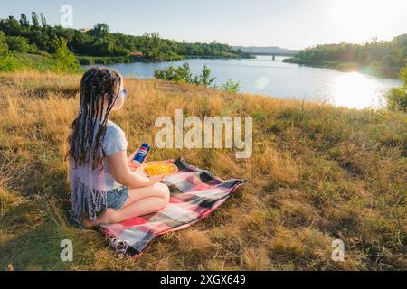 Krivoy Rog, Ucraina, 26 giugno 2024: Ragazza seduta sulla riva del fiume e tiene in mano una lattina di Pepsi Cola e un piatto di cartone con patatine fritte Foto Stock