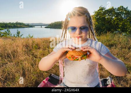 Una ragazza con gli occhiali blu si siede sulla riva del fiume e tiene un hamburger in mano. Soleggiata giornata estiva in campagna. Picnic all'aperto Foto Stock