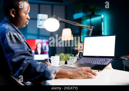 Lavoratrice libera professionista che utilizza la tecnologia mentre lavora a casa con un notebook digitale dotato di schermo bianco isolato. Donna nera che guarda un personal computer che visualizza un modello di mockup vuoto. Foto Stock