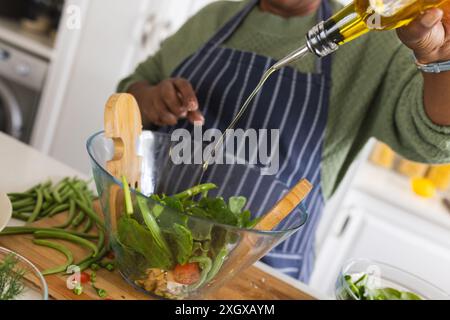 Donna afroamericana anziana che prepara insalata e versa olio in cucina a casa. Vita domestica, pensione, cibo e stile di vita, inalterati. Foto Stock