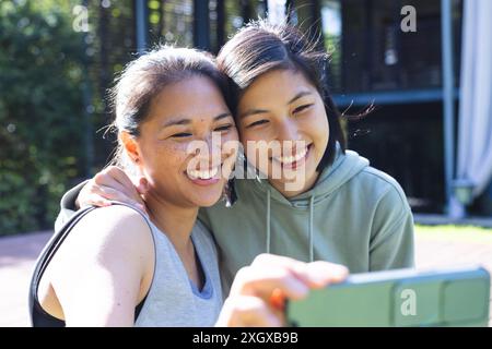 Amiche asiatiche felici che usano lo smartphone e si abbracciano in giardino a casa. Fitness, esercizio fisico, vita domestica, amicizia, attività e stile di vita, una Foto Stock