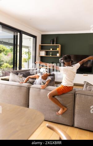 Fratello e sorella birazziali adolescenti apprezzano la realtà virtuale a casa. Sono immersi in un'esperienza di realtà virtuale in un accogliente salotto. Foto Stock