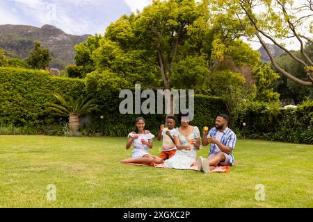 Una giovane famiglia birazziale ama fare un picnic all'aperto. Condividono un momento di gioia con gelati rinfrescanti in un giardino lussureggiante. Foto Stock