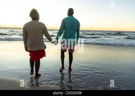 Una coppia afroamericana senior gode di una spiaggia tranquilla al tramonto, con spazio per fotocopie. Condividono un momento tranquillo camminando lungo il litorale, riflettendo Foto Stock