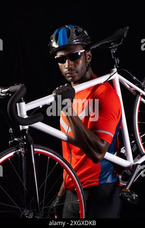 Un uomo afroamericano posa con la sua bicicletta su uno sfondo nero, con spazio di copia. È vestito con l'attrezzatura da ciclismo, pronto per un giro notturno o una gara. Foto Stock