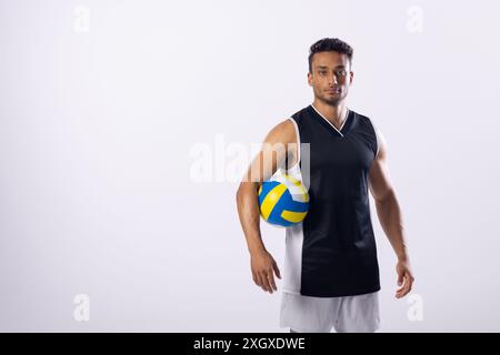 Il giovane uomo birazziale posa con sicurezza con una pallavolo su sfondo bianco, con spazio di copia. Il suo abbigliamento atletico suggerisce la passione per lo sport e per la a Foto Stock
