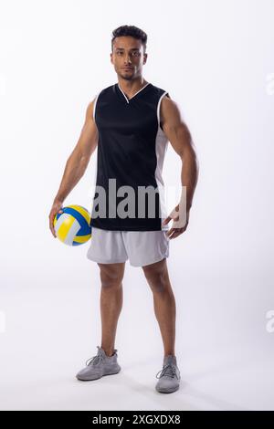 Il giovane uomo birazziale si pone con sicurezza nell'abbigliamento sportivo, tenendo una pallavolo su uno sfondo bianco. Il suo abbigliamento atletico suggerisce una preparazione per una partita o. Foto Stock