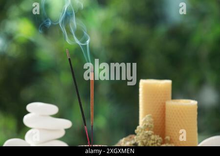 Bastoncini di incenso che si muovono vicino a pietre, candele e fiori secchi su sfondo verde sfocato Foto Stock