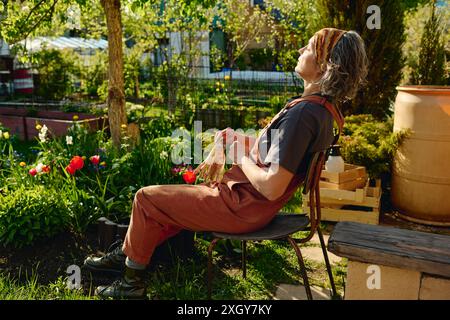 Vista laterale di una donna matura giardiniera con t-shirt grigia, tute marroni e archetto seduto su una sedia in giardino e godendosi la luce del sole Foto Stock