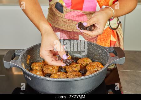 Una donna mette le prugne secche in una padella con cotolette di carne e condimenti sul piano cottura elettrico della sua cucina di casa. Foto Stock