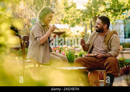 Allegri giardinieri maturi e maschi che chiacchierano e mangiavano patate cotte mentre sedevano tra alberi e piante verdi Foto Stock