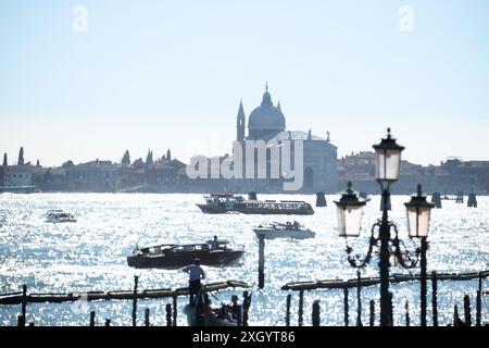 Barche che attraversano la scintillante distesa d'acqua tra Riva degli Schiavoni e la Chiesa del Santissimo Redentore incorniciata da una lampada e da un cielo azzurro soffice Foto Stock