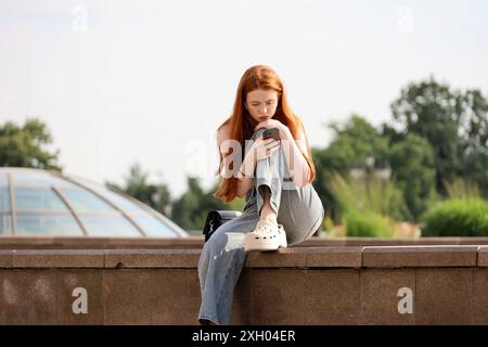 Redhead girl seduta con lo smartphone in strada. Utilizzo del telefono cellulare in città estiva Foto Stock