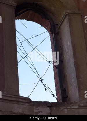 Finestra ad arco rotta in un edificio abbandonato con fili esposti contro un cielo limpido, che ritrae decadimento e abbandono. Foto Stock