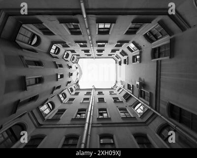 Immagine in bianco e nero di uno stretto cortile in un vecchio edificio visto dal basso, creando un effetto tunnel con finestre. Foto Stock